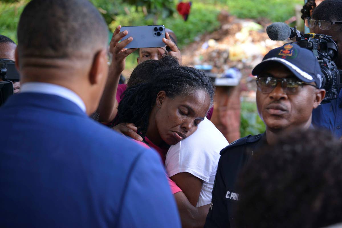 Prime Minister Dr Andrew Holness looks on as two grieving family members embrace each other along the Commodore Road in Linstead, St Catherine.