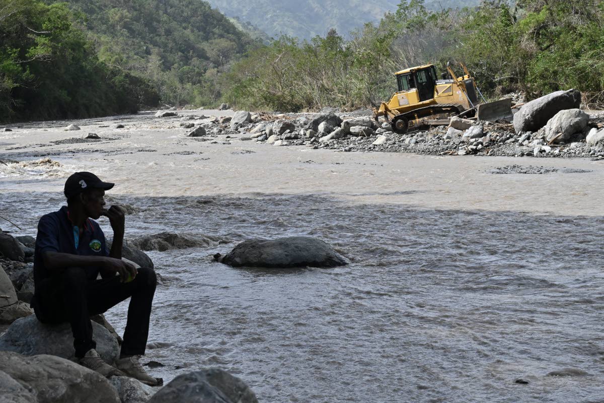 A resident sits on the bank of the Yallahs River on Monday as a tractor driver tries to redirect the course of the raging river so people can cross the former fording after the four-lane bridge collapsed last week as Hurricane Melissa passed.