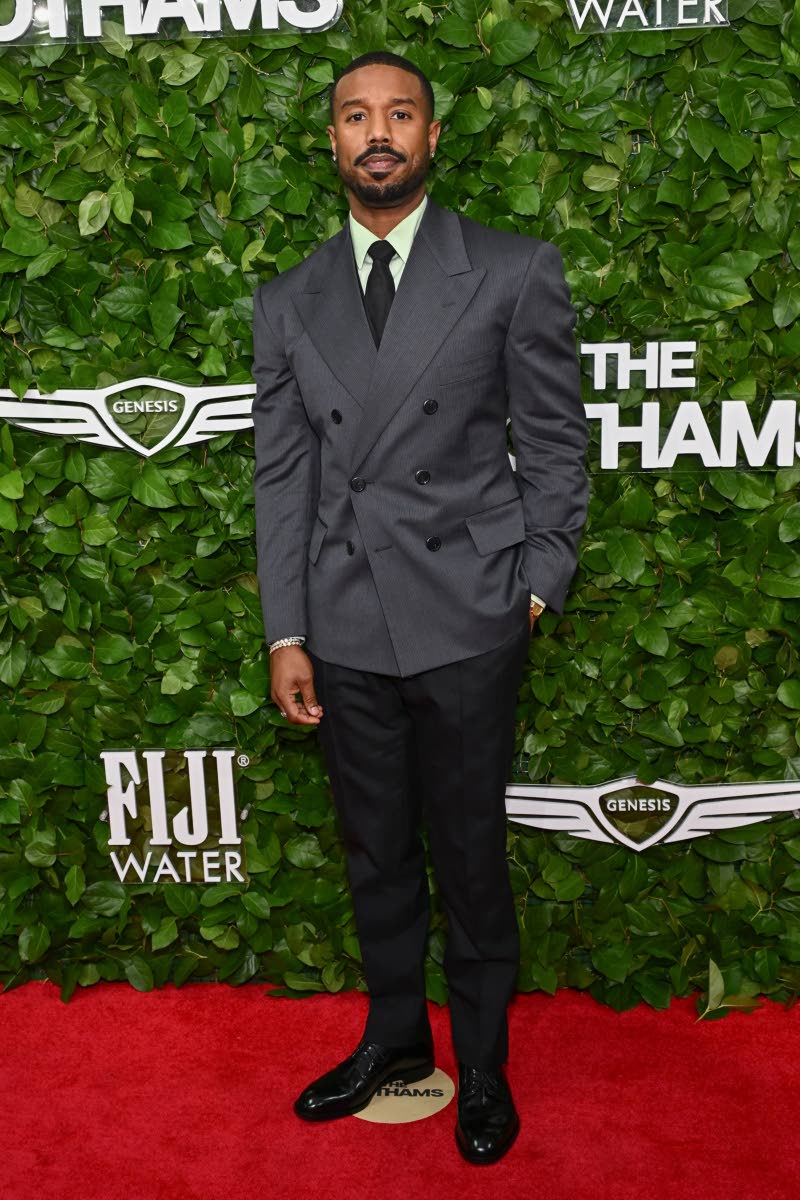Michael B. Jordan dons a Prada dark slate grey double-breasted blazer, soft green poplin shirt, and satin black tie paired with black brushed leather lace-ups.