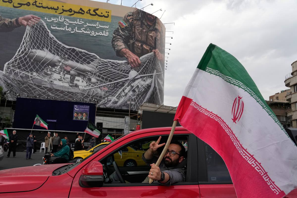A man flashes a victory sign as he carries an Iranian flag in front of an anti-US billboard depicting the American aircrafts into the Iranian armed forces fishing net with signs that read in Farsi: “The Strait of Hormuz will remain closed. The entire Per