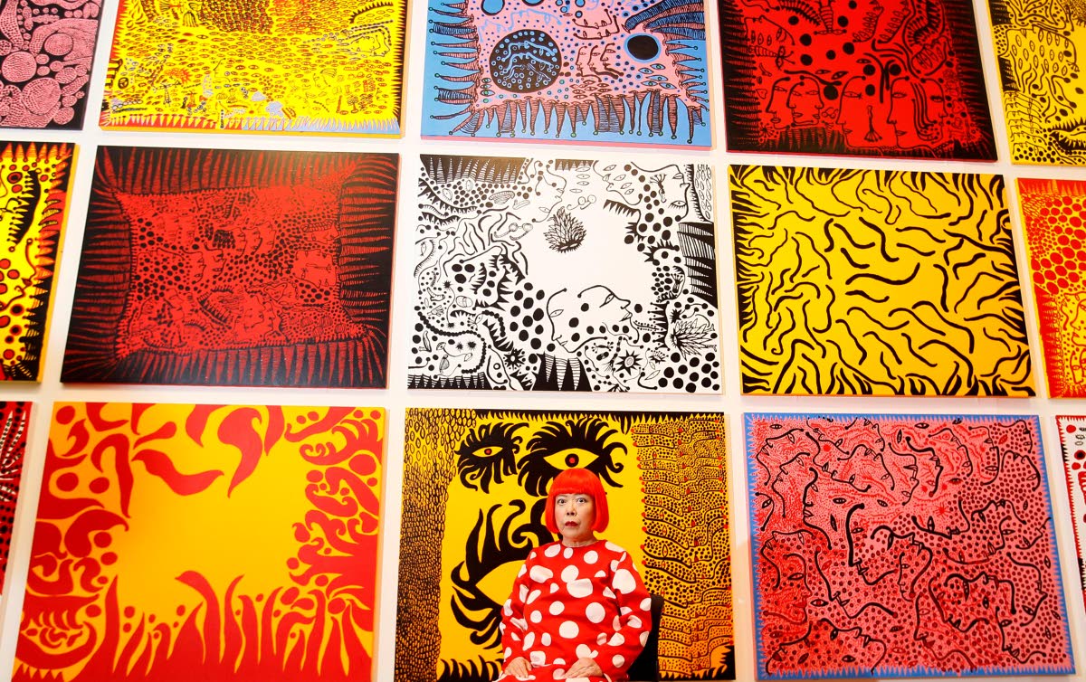 Japanese artist Yayoi Kusama poses for photographers in front of her painting ‘Love Arrives at the Earth Carrying with it a Tale of the Cosmos’ (centre bottom), and other paintings at the Tate Modern in London on February 7, 2012.