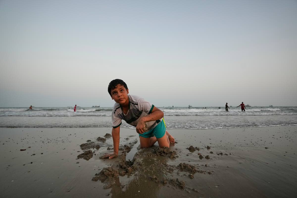 A boy plays in wet sand as others swim and commercial vessels lie anchored in the Strait of Hormuz off Bandar Abbas, Iran.