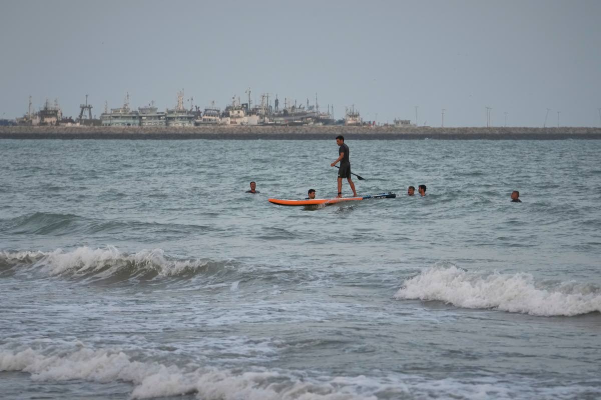 A man paddleboards among swimmers in the Strait of Hormuz, with boats docked at a harbor in Bandar Abbas, Iran.