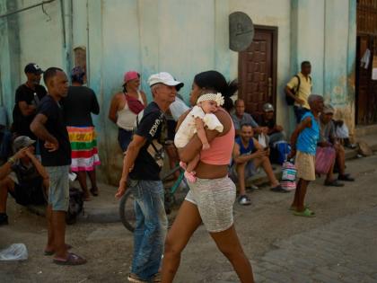 A woman walks with a baby in her arms past people lined up to buy bread during a blackout in Havana, Cuba,