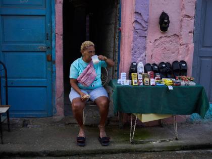 A vendor having breakfast sits by her table holding various products, from cigarettes to sandals, in Havana, Cuba.
