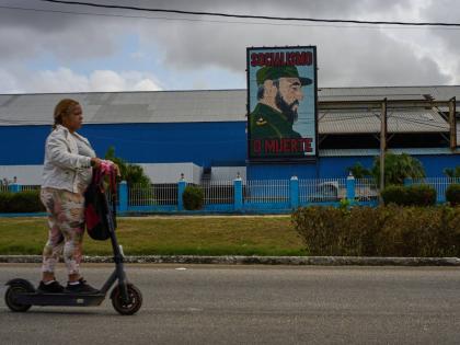 A woman rides an electric scooter past a factory displaying an image depicting the late Cuban leader Fidel Castro, bearing the words "Socialism or Death", in Havana, Cuba on March 19, 2026. (AP Photo/Ramon Espinosa)