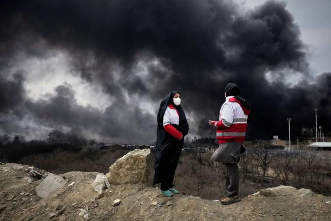 Two women from the Iranian Red Crescent Society stand as a thick plume of smoke from a US-Israeli strike on an oil storage facility rises in the sky in Tehran.