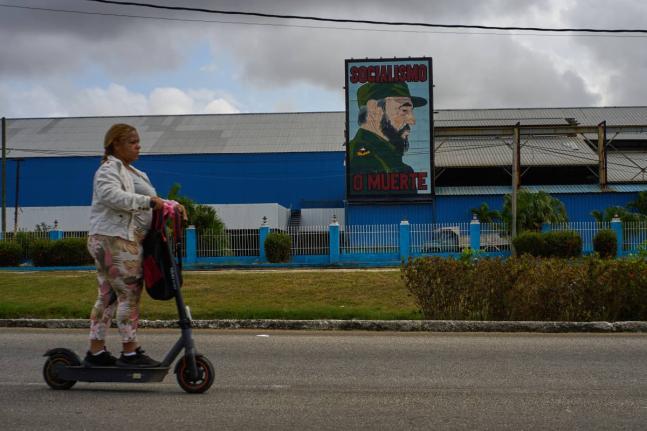 A woman rides an electric scooter past a factory displaying an image depicting the late Cuban leader Fidel Castro, bearing the words "Socialism or Death", in Havana, Cuba on March 19, 2026. (AP Photo/Ramon Espinosa)