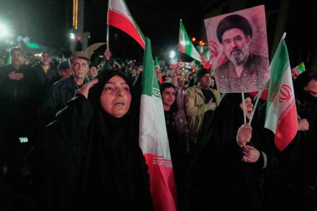 A woman salutes while singing the national anthem as another holds a picture of Iranian Supreme Leader Ayatollah Mojtaba Khamenei during a pro-government gathering in a square in western Tehran, Iran on March 25, 2026. (AP Photo/Vahid Salemi)