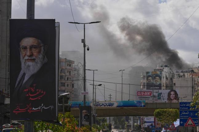A portrait of Iran's late Supreme Leader Ayatollah Ali Khamenei, left, is seen, as smoke rises following an Israeli airstrike in Dahiyeh, Beirut's southern suburbs, Lebanon on March 30, 2026. (AP Photo/Hassan Ammar)