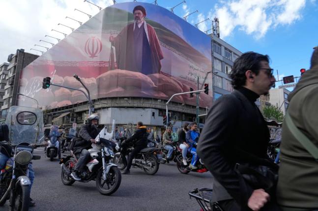 People drive their motorbikes past a billboard that shows a graphic depicting Iran's Supreme Leader Ayatollah Mojtaba Khamenei in downtown Tehran, Iran in April 6, 2026. (AP Photo/Vahid Salemi)