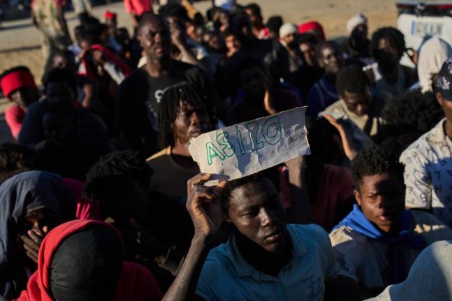 A migrant who crossed from Morocco into Spain holds a sign reading, in Spanish, “asylum” as he lines up for food distribution on a beach in the Spanish enclave of Ceuta, Monday, August 3, 2026. 