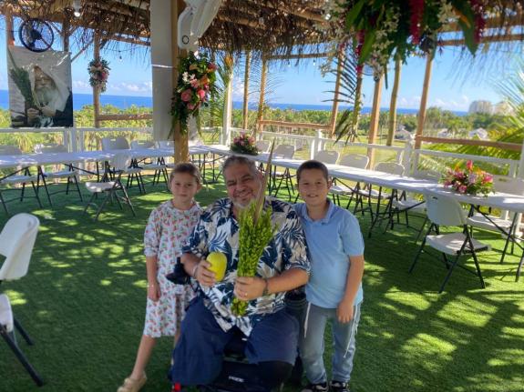 Chava and Moshe Raskin shake the lulav with Gavriel Abigzer, who lost both of his legs. Even while on vacation in Jamaica, Gavriel made the special effort to fulfill the mitzvah of lulav and etrog.